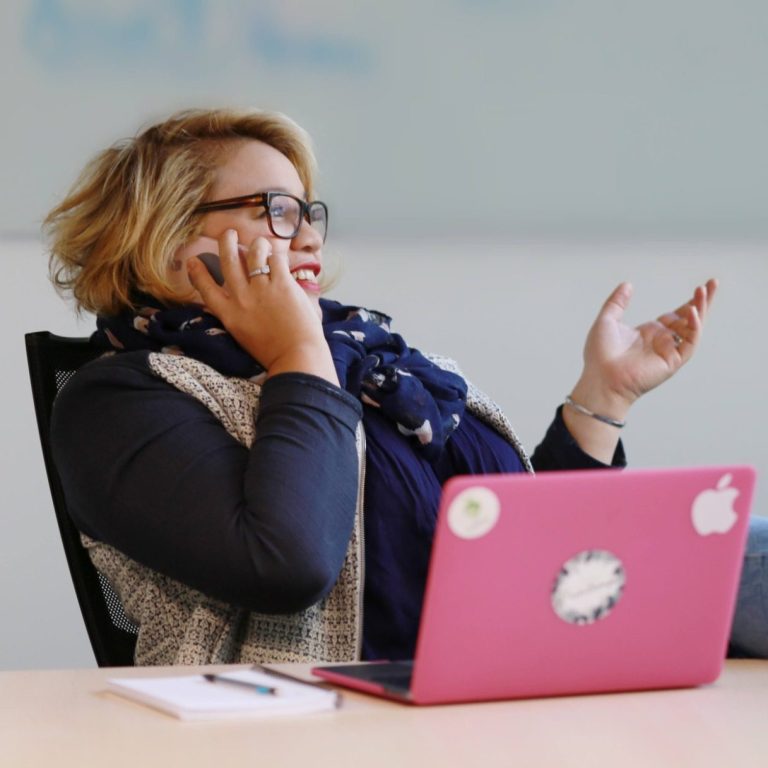 FAQ : une femme au téléphone devant son ordinateur portable, la main levée d'un air interrogatif