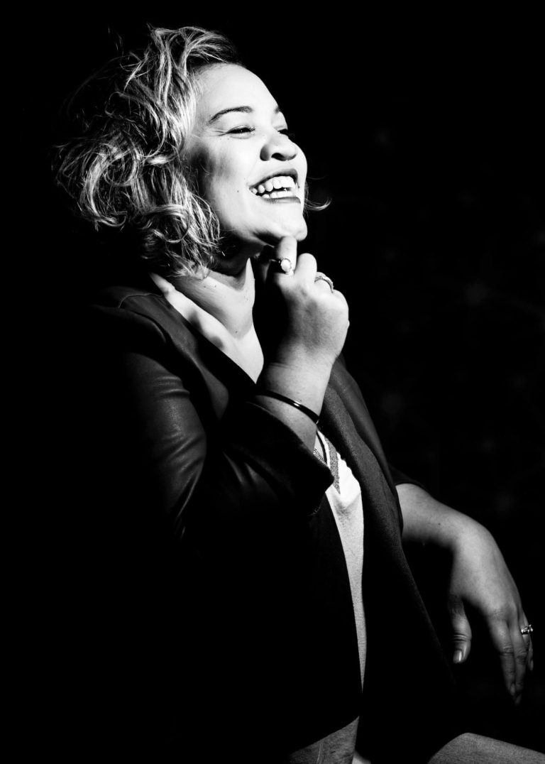 Femme souriante avec des cheveux bouclés, portant un blazer noir sur fond sombre.