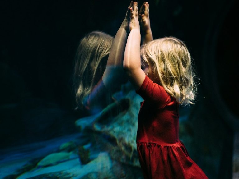 Une petite fille blonde en robe rouge se tient devant un miroir, mains levées.